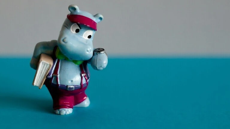Plava figurica nilskog konja iz Kinder jaja iz 80-ih, stoji na plavoj podlozi i drži knjigu.